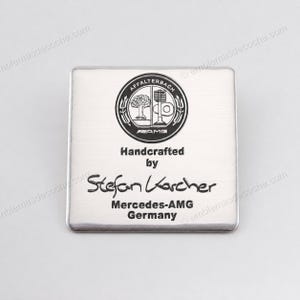 Puede incluir: Una placa cuadrada de metal cepillado con un emblema circular negro y plateado de AMG Affalterbach en la parte superior. Debajo del emblema, el texto dice Handcrafted by Stefan Karcher, seguido de Mercedes-AMG Germany en letras negras.