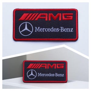 Può includere: Toppa rettangolare nera con testo e logo ricamati in rosso e bianco. Il testo rosso recita "AMG" sopra un logo Mercedes-Benz bianco e le parole "Mercedes-Benz". La toppa ha un bordo rosso.