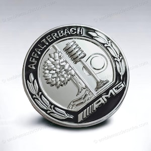 Könnte beinhalten: Ein silbernes und schwarzes AMG-Emblem mit dem Text "AFFALTERBACH" am oberen Rand. Das Emblem zeigt einen Baum, ein Ventil und das AMG-Logo. Das Emblem ist kreisförmig und hat eine glänzende, metallische Oberfläche.