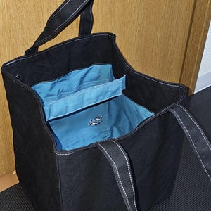 May include: Black tote bag with white stitching on the handles and seams. The interior is a bright blue with pockets. A small emblem is visible on the inside. The bag is open, revealing the interior.