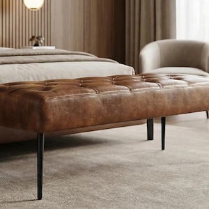 May include: A brown leather bench with a tufted design and black metal legs. The bench is rectangular and appears to be in a bedroom setting, with a bed and chairs in the background. The bench is approximately 48 inches long.