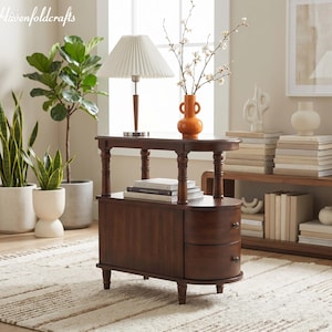 May include: A dark brown wooden side table with two drawers and a curved front. A white pleated lampshade sits on top, along with an orange vase with white flowers. The table is on a patterned rug.