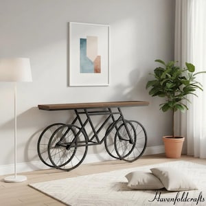 May include: A unique console table with a bicycle wheel design. The table features a dark brown wooden top supported by a black metal frame shaped like a bicycle. The wheels are visible, adding to the artistic design. The table is placed against a light gray wall.