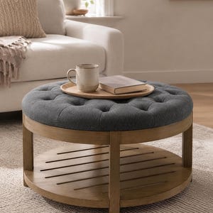 May include: A round ottoman with a dark gray tufted fabric top and a light brown wooden frame. The base features a slatted circular shelf. A light brown wooden tray sits on the top, holding a ceramic mug and a closed book with a light cover.