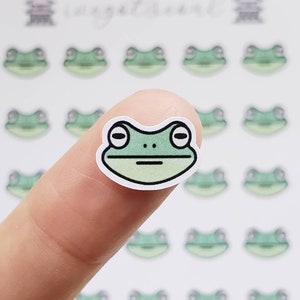 May include: A sheet of stickers featuring multiple green frog faces. The stickers are on a white background. One sticker is being held by a finger.