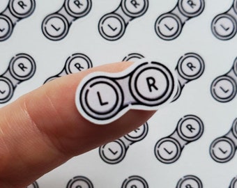 May include: A sheet of contact lens stickers with a white background and black outlines. The stickers are in the shape of contact lens cases with the letters "L" and "R" printed on them. The stickers are arranged in a grid pattern.