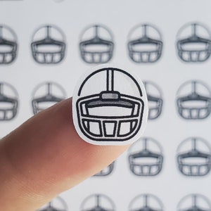 May include: A sheet of white sticker paper with multiple black and white football helmet stickers. One sticker is being held by a finger.