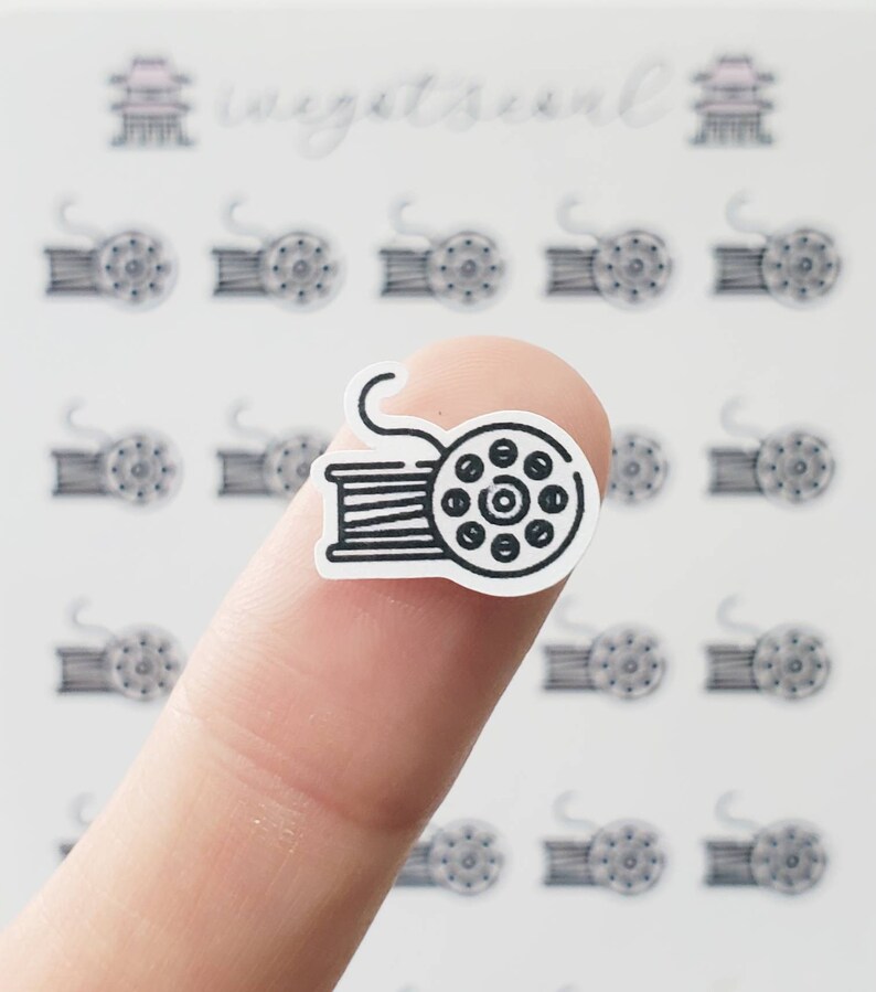 May include: A black and white sticker of a spool of thread with a string attached. The sticker is on a finger.