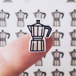 May include: A small, white sticker with a black and white illustration of a stovetop espresso maker, held between fingers. The background shows a pattern of similar espresso maker illustrations.