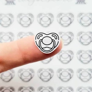 May include: A sheet of white sticker paper with multiple black and white pacifier outline stickers. One sticker is being held by a finger.