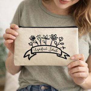 May include: Beige canvas zippered pouch with a black floral design and the text "Useful Shit" on a banner. The pouch is rectangular and held by a person. The background is blurred.