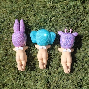 Puede incluir: Tres figuras caprichosas con cabezas de animales enjoyadas: un conejo morado, un elefante azul y una jirafa morada. Cada figura tiene un cuerpo color piel y un cuello blanco. Las figuras se exhiben sobre hierba verde.