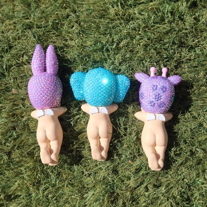 Puede incluir: Tres figuras caprichosas con cabezas de animales enjoyadas: un conejo morado, un elefante azul y una jirafa morada. Cada figura tiene un cuerpo color piel y un cuello blanco. Las figuras se exhiben sobre hierba verde.