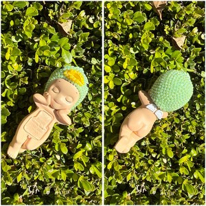 Puede incluir: Dos figuras de beb&eacute; de cer&aacute;mica, una con un sombrero verde y amarillo, la otra con una cabeza de cuentas verdes. Ambos est&aacute;n acostados sobre un lecho de hojas verdes. Las figuras tienen los ojos cerrados y est&aacute;n en una pose relajada.