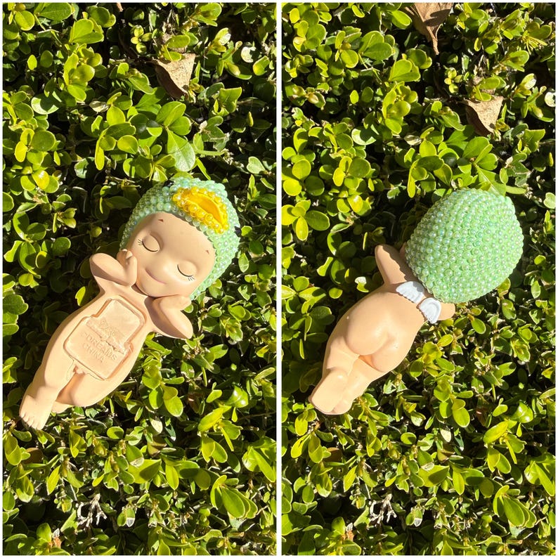 Puede incluir: Dos figuras de beb&eacute; de cer&aacute;mica, una con un sombrero verde y amarillo, la otra con una cabeza de cuentas verdes. Ambos est&aacute;n acostados sobre un lecho de hojas verdes. Las figuras tienen los ojos cerrados y est&aacute;n en una pose relajada.