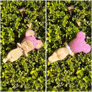 Puede incluir: Dos figuras con forma de beb&eacute;, una tumbada boca arriba y otra boca abajo, ambas con un gorro rosa con orejas de rat&oacute;n enjoyado. Las figuras son de color beige, con los ojos cerrados y una expresi&oacute;n serena. Descansan sobre hojas verdes.