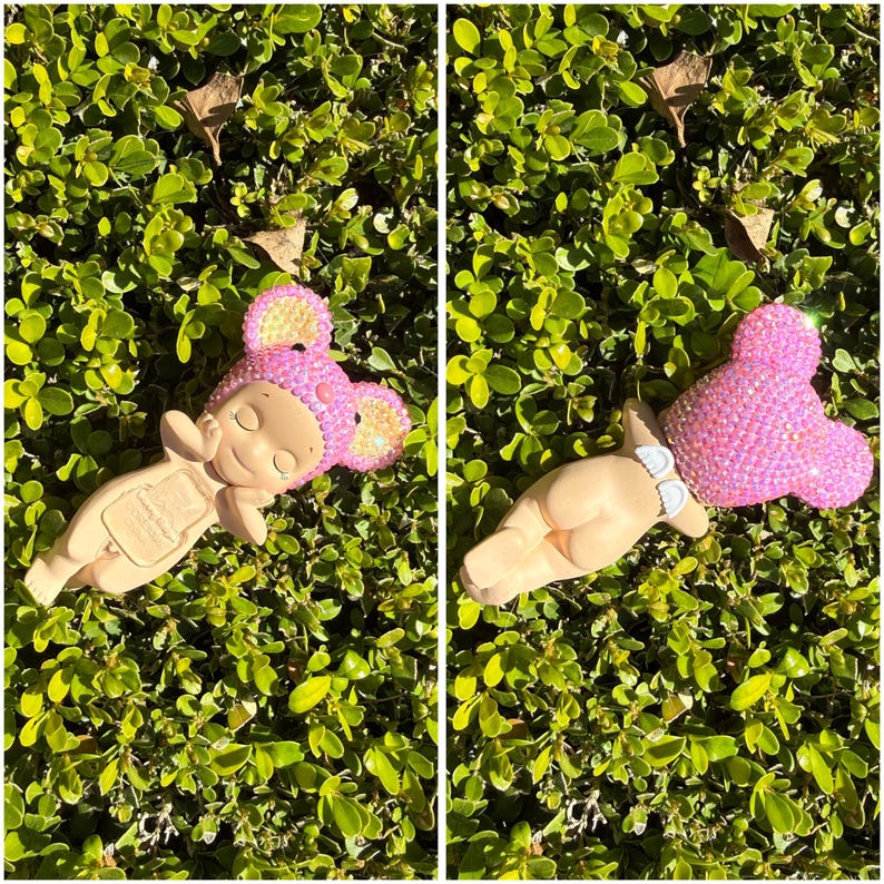 Puede incluir: Dos figuras con forma de beb&eacute;, una tumbada boca arriba y otra boca abajo, ambas con un gorro rosa con orejas de rat&oacute;n enjoyado. Las figuras son de color beige, con los ojos cerrados y una expresi&oacute;n serena. Descansan sobre hojas verdes.