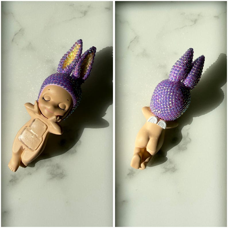 Puede incluir: Una figura de un beb&eacute; con un gorro de conejito morado enjoyado y orejas a juego. El beb&eacute; est&aacute; desnudo con los brazos extendidos. El gorro de conejito tiene detalles amarillos. La figura est&aacute; en una pose relajada.