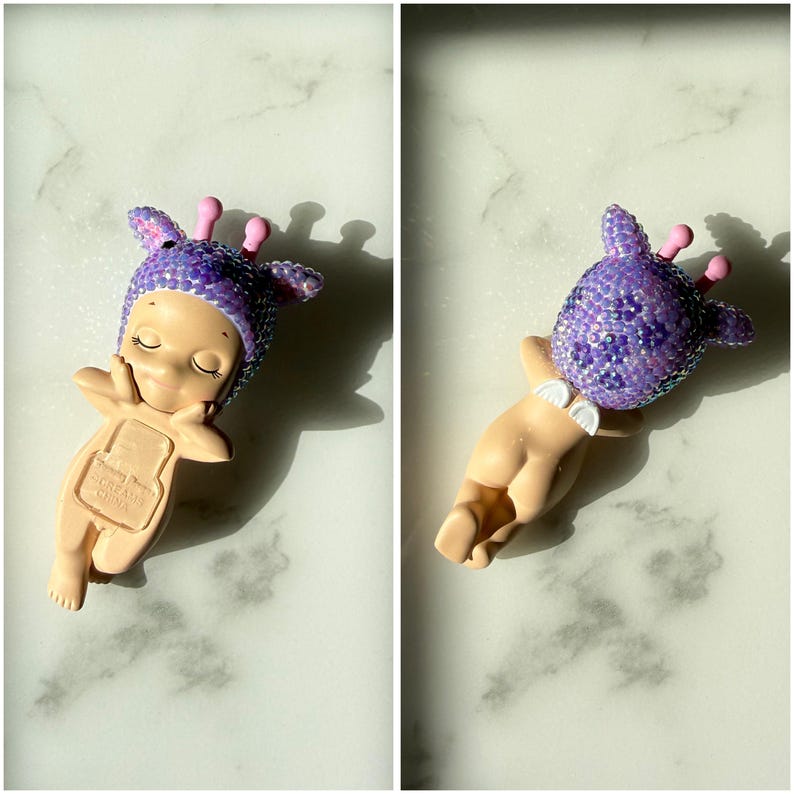 Puede incluir: Peque&ntilde;a figura de beb&eacute; de color beige con un gorro morado con lentejuelas y detalles rosas. El beb&eacute; est&aacute; en una pose relajada, con las manos cerca de la cara. La figura tiene peque&ntilde;as alas blancas en la espalda.