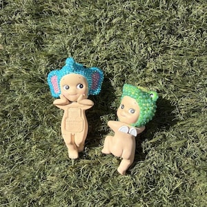 Puede incluir: Dos peque&ntilde;as figuras sobre hierba verde. Una figura tiene orejas de elefante azules y detalles en rosa. La otra figura tiene un sombrero de rana verde y peque&ntilde;as alas blancas. Ambas figuras tienen caras sonrientes y son de color marr&oacute;n claro.