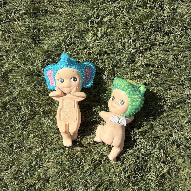 Puede incluir: Dos peque&ntilde;as figuras sobre hierba verde. Una figura tiene orejas de elefante azules y detalles en rosa. La otra figura tiene un sombrero de rana verde y peque&ntilde;as alas blancas. Ambas figuras tienen caras sonrientes y son de color marr&oacute;n claro.