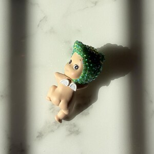 Puede incluir: Peque&ntilde;a figura de color carne con un tocado verde enjoyado y peque&ntilde;as alas blancas. La figura tiene ojos grandes y est&aacute; sobre una superficie de m&aacute;rmol blanco. La sombra de la figura se proyecta hacia la derecha.