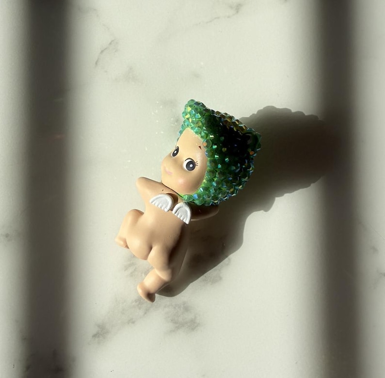 Puede incluir: Peque&ntilde;a figura de color carne con un tocado verde enjoyado y peque&ntilde;as alas blancas. La figura tiene ojos grandes y est&aacute; sobre una superficie de m&aacute;rmol blanco. La sombra de la figura se proyecta hacia la derecha.