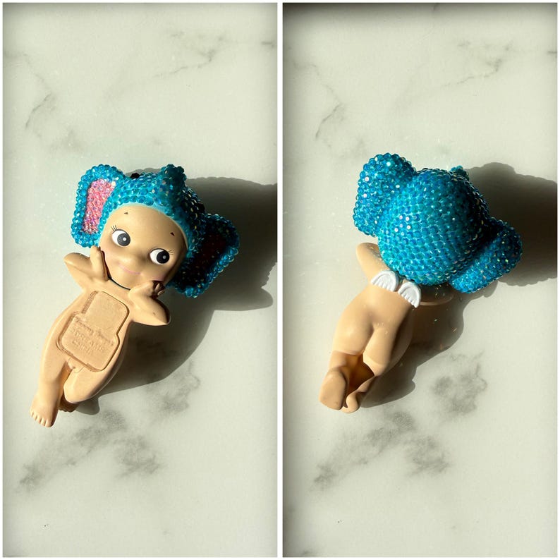 Puede incluir: Figura de un beb&eacute; con un sombrero de elefante azul con detalles en rosa. El beb&eacute; tiene la piel clara y est&aacute; en posici&oacute;n reclinada. El sombrero est&aacute; cubierto de peque&ntilde;os adornos brillantes. La figura tiene un dise&ntilde;o caprichoso y juguet&oacute;n.