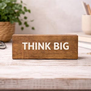 May include: A wooden block with the words "THINK BIG" in white lettering. The block is on a white wooden surface. In the background are a potted plant, eyeglasses, a mug with pencils, and books.