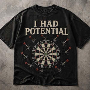 Può includere: T-shirt nero con la scritta "I HAD POTENTIAL" sopra un bersaglio per le freccette. Il bersaglio è circondato da freccette. La maglietta è di un colore scuro e sbiadito.