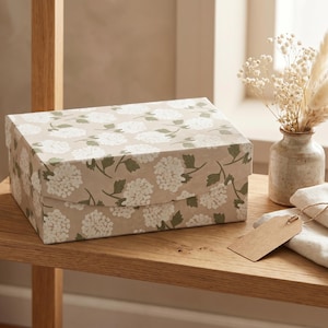 May include: A rectangular gift box with a floral pattern of white flowers and green leaves on a tan background. The box is on a wooden shelf, next to a small vase with dried flowers and a folded cloth with a tag.