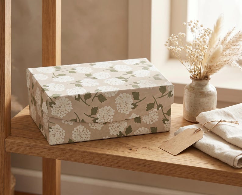 May include: A rectangular gift box with a floral pattern of white flowers and green leaves on a tan background. The box is on a wooden shelf, next to a small vase with dried flowers and a folded cloth with a tag.