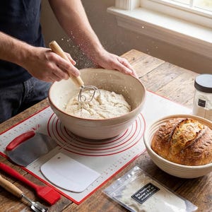May include: A person mixing dough in a beige ceramic bowl with a wooden handled whisk. A red and white silicone mat with measurement markings, a loaf of bread in a woven basket, and baking tools are on a wooden table. A bag of dry sourdough starter is visible.