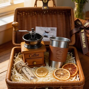 May include: A coffee-themed gift basket with a wooden coffee grinder, a stainless steel tumbler with a leather sleeve, and dried orange slices. The basket also contains a small glass bottle with the text "Thank you".