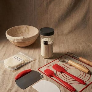 May include: A bread-making kit with a banneton basket, a jar of flour, and various tools. Includes a dough scraper, a pastry brush, a silicone mat, and a lame. The kit also contains a packet of sourdough starter.