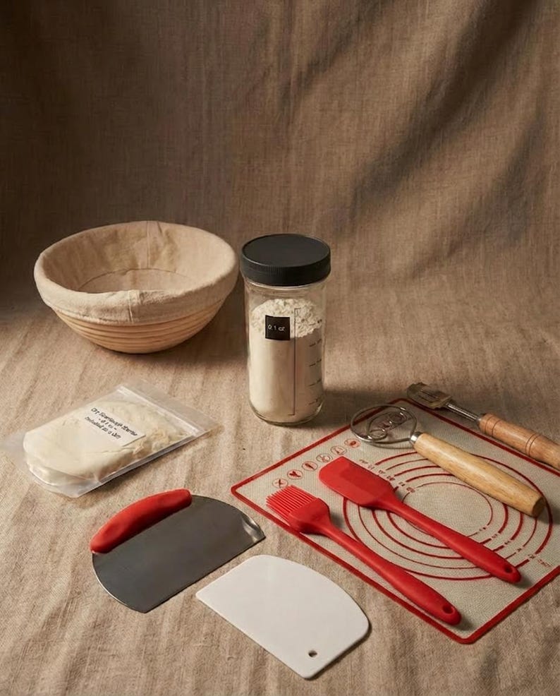 May include: A bread-making kit with a banneton basket, a jar of flour, and various tools. Includes a dough scraper, a pastry brush, a silicone mat, and a lame. The kit also contains a packet of sourdough starter.