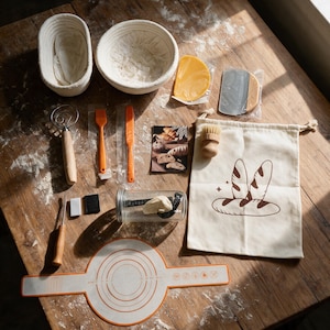 May include: A sourdough bread baking kit including two oval and round proofing baskets, a silicone mat with measurement markings, a dough whisk, orange silicone tools, a metal scraper, a wooden brush, and a canvas drawstring bag with a bread graphic.