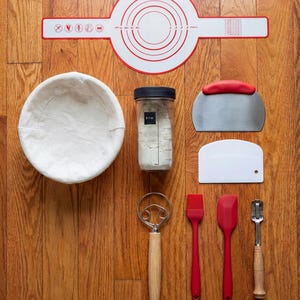 May include: A collection of baking tools arranged on a wooden surface. Includes a red and white silicone mat, a white linen-lined proofing basket, a jar of flour, a dough scraper, a bench scraper, a whisk, a brush, a spatula, and a lame.