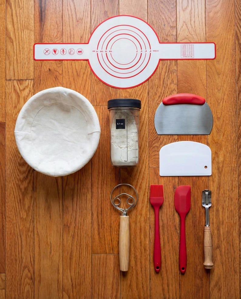 May include: A collection of baking tools arranged on a wooden surface. Includes a red and white silicone mat, a white linen-lined proofing basket, a jar of flour, a dough scraper, a bench scraper, a whisk, a brush, a spatula, and a lame.
