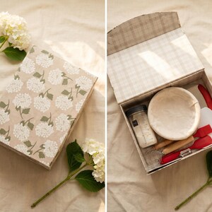 May include: A beige box with a floral pattern, containing baking tools. Inside, a round proofing basket, a jar, and red and white utensils are visible. The box is open, revealing the contents, and is placed on a light-colored surface.