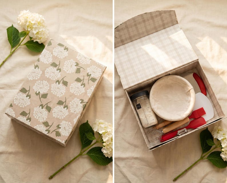 May include: A beige box with a floral pattern, containing baking tools. Inside, a round proofing basket, a jar, and red and white utensils are visible. The box is open, revealing the contents, and is placed on a light-colored surface.