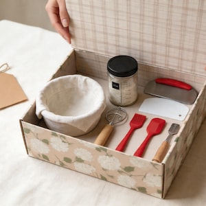 May include: A baking kit in a floral patterned box. The kit includes a proofing basket, jar, dough scraper, whisk, and red silicone tools. A blank tag with twine is on the left. The box is open, revealing the contents.