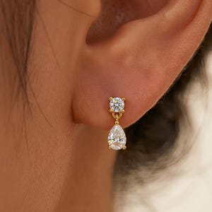 May include: A gold-colored earring with a round, clear gemstone stud and a dangling pear-shaped, clear gemstone. The earring is worn on a person's ear.