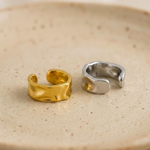 May include: Two ear cuffs, one in a polished gold tone and one in a polished silver tone. Both pieces feature an open, irregular, and wavy band design with a smooth, reflective surface finish, showcasing a distinct organic shape.