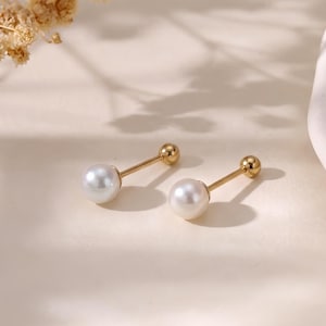 May include: A pair of gold-tone barbell earrings, each featuring a smooth, round white pearl at one end and a small gold-tone ball at the other. The earrings have a straight metal post connecting the pearl to the back closure, creating a simple design.