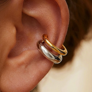 Könnte beinhalten: Nahaufnahme eines Ohrs mit zwei Ohrklemmen. Eine ist goldfarben, die andere silberfarben. Die Ohrklemmen sind glatt und abgerundet und schaffen eine moderne, minimalistische Ästhetik. Der Schmuck wird am oberen Ohr getragen.