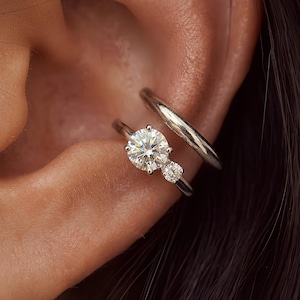 May include: A silver-toned ear cuff and a silver-toned ring featuring a large round brilliant-cut stone and a smaller round accent stone. The jewelry is positioned on the outer cartilage and the inner conch area of the ear, showcasing a metallic finish.