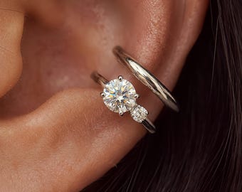 May include: A silver-toned ear cuff and a silver-toned ring featuring a large round brilliant-cut stone and a smaller round accent stone. The jewelry is positioned on the outer cartilage and the inner conch area of the ear, showcasing a metallic finish.