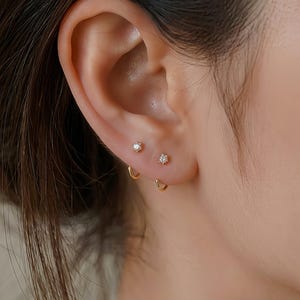 May include: Two small gold stud earrings featuring clear, round-cut stones set in a six-prong design, positioned side-by-side on the earlobe. Below these studs, two thin, gold hoop earrings are inserted into the earlobe piercings.