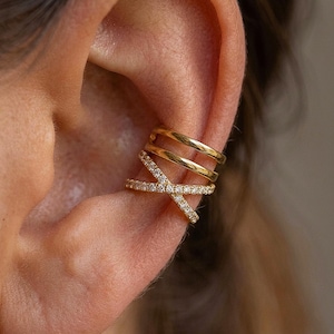 May include: A gold ear cuff featuring three bands and a crisscross pattern embellished with small, sparkling stones. The jewellery is worn on the ear and appears to be made of gold.